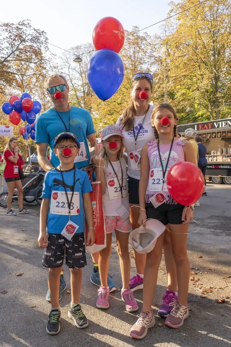 Eine Familie posiert mit roten Nasen und Luftballons bei einem Rennevent, mit Bäumen und einem Imbisswagen im Hintergrund.
