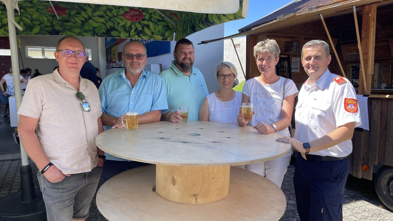 Eine Gruppe von sechs Erwachsenen, vier Männer und zwei Frauen, lächeln und posieren für ein Foto mit Gläsern Bier. Sie stehen um einen runden Holztisch mit einem Fasssockel herum, unter einem Regenschirm mit einem roten Apfeldesign.