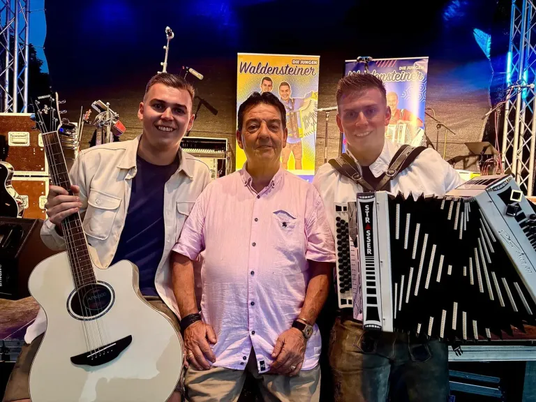 Drei Männer stehen lächelnd für ein Foto zusammen. Der Mann auf der linken Seite hält eine Gitarre. Der mittlere Mann trägt ein rosa Hemd und eine Uhr. Der rechte Mann spielt Akkordeon. Hinter ihnen befinden sich Banner mit Text und Bildern.