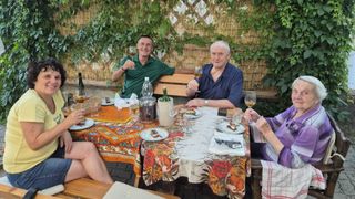 Vier Personen sitzen an einem Tisch im Freien, jede mit einem Weinglas in der Hand. Ein Tischtuch mit Blumenmustern liegt auf dem Tisch. Flaschen und Teller stehen auf dem Tisch. Eine Pflanze wächst an der Wand hinter ihnen.