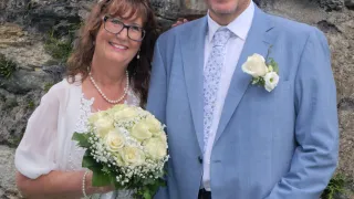 Ein Mann und eine Frau in formeller Kleidung stehen lächelnd vor einer Steinmauer. Die Frau hält einen Strauß weißer Blumen.