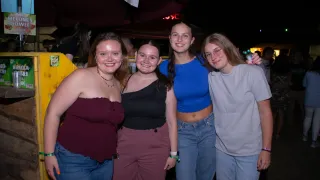 Vier Frauen lächeln und posieren für ein Foto bei Nacht. Die Frau auf der linken Seite trägt ein Weinrotoberteil, die daneben ein schwarzes Top, die in der Mitte ein blaues Oberteil und die auf der rechten Seite ein graues Top.