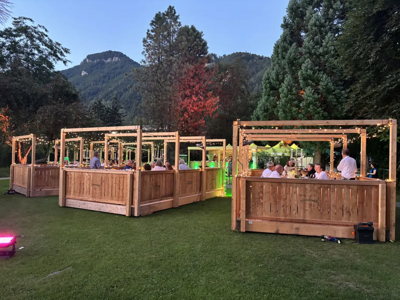 Ein Außenbereich mit hölzernen Sitznischen und Lichterketten, umgeben von Bäumen und Bergen, mit sitzenden und stehenden Menschen.