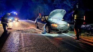 Ein beschädigtes Auto steht bei Nacht am Straßenrand. Die Motorhaube ist offen, und die Front ist demoliert. Eine Person steht in der Nähe.