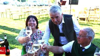 Drei Personen versammeln sich draußen und halten Weingläser. Eine Frau lächelt und schaut in die Kamera. Ein Mann im Vest lächelt und hält ein Glas. Ein anderer Mann schaut in die Kamera. Ein Tisch mit einer Flasche ist in der Nähe.
