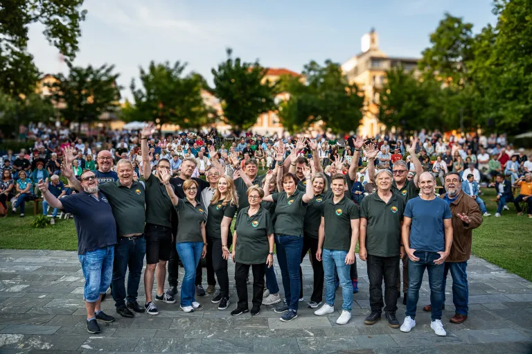 Eine große Gruppe von Menschen in passenden grünen Shirts und Jeans steht in einem Park mit Bäumen und Gebäuden im Hintergrund. Sie heben ihre Hände und lächeln für ein Foto.