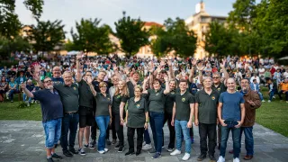 Eine große Gruppe von Menschen in passenden grünen Shirts und Jeans steht in einem Park mit Bäumen und Gebäuden im Hintergrund. Sie heben ihre Hände und lächeln für ein Foto.