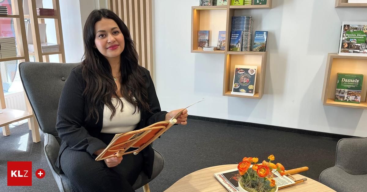Eine Frau in einem schwarzen Blazer und einem weißen Oberteil sitzt in einem Stuhl, hält ein offenes Buch und lächelt. Hinter ihr ist eine weiße Wand mit Bücherregalen zu sehen. Auf einem Tisch stehen Blumen in einem Topf.