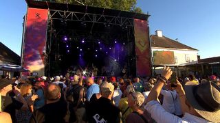 Ein lebhaftes Open-Air-Musikfestival mit einer großen Menschenmenge, die die Darbietung auf der Bühne unter einer bunten Lichtshow und einem klaren blauen Himmel genießt.