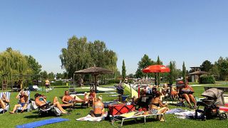 Eine Gruppe von Menschen entspannt sich auf dem Rasen in einem Park. Einige tragen Badeanzüge, während andere unter Sonnenschirmen sitzen. Bäume und ein Schwimmbecken sind im Hintergrund zu sehen.