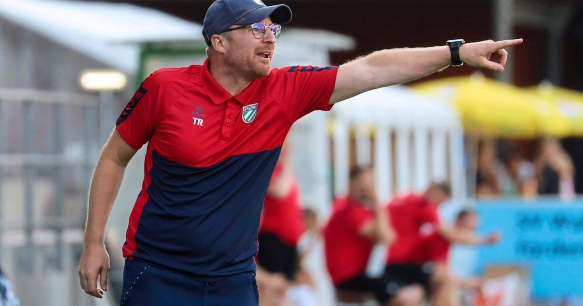 Ein männlicher Trainer in einem rot-blauen Shirt, mit den Buchstaben TR auf der linken Seite, gestikuliert und gibt seinen Spielern Anweisungen. Die Spieler sind im Hintergrund verschwommen.