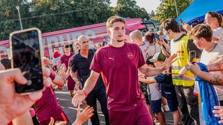 Ein Mann in einem weinroten Trikot begrüßt Fans vor einem roten Bus, mit Menschen und Bäumen im Hintergrund.