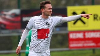 Ein Fußballspieler in einem weißen Trikot mit einem roten Quadratdesign auf der Vorderseite, der seinen rechten Arm ausstreckt und zur Seite schaut.