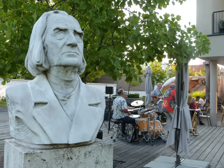 Eine Büste eines Mannes mit langen Haaren steht auf einem Podest. Hinter der Büste spielt eine Gruppe von Menschen Musikinstrumente auf einer Holzterrasse.