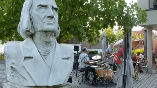 Eine Büste eines Mannes mit langen Haaren steht auf einem Podest. Hinter der Büste spielt eine Gruppe von Menschen Musikinstrumente auf einer Holzterrasse.