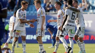 Fußballspieler in weißen Trikots stehen auf einem Feld, einer davon hockt sich hin. Der vordere Spieler hat ein Logo auf seinem Trikot. Ein weiterer Spieler hinter ihm trägt Turnschuhe.