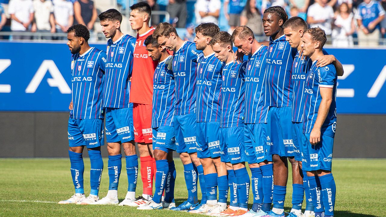 Eine Fußballmannschaft in blauen Trikots steht auf dem Feld, den Kopf gesenkt als Zeichen des Respekts. Die blauen Trikots der Mannschaft sind mit weißen Streifen und verschiedenen Sponsorenlogos versehen. Der Spieler in Rot sticht hervor, während verschwommene Zuschauer im Hintergrund zu sehen sind.