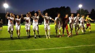 Eine Gruppe männlicher Fußballspieler, die auf einem Rasenfeld mit einer weißen Linie feiern, tragen Trikots mit Nummern und Logos.