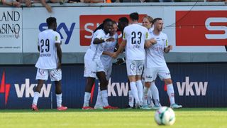 Fußballspieler in weißen Trikots feiern auf dem Spielfeld. Spieler 29 steht abseits, während sich die anderen um Spieler 23 scharen. Ein Fußball liegt auf dem Boden.