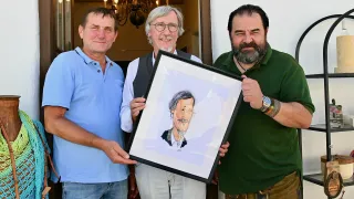 Drei Männer halten eine gerahmte Karikatur eines lächelnden Mannes mit Schnurrbart. Sie stehen in einem Raum, hinter ihnen hängt eine Lampe.