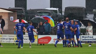 Eine Fußballmannschaft feiert in der Regen einen Treffer, in blauen Trikots mit den Nummern 11, 9, 20, 18 und 24. Zuschauer im Hintergrund halten Regenschirme. Das Spielfeld hat ein Banner mit dem Text 'Markisen' und einem Logo.
