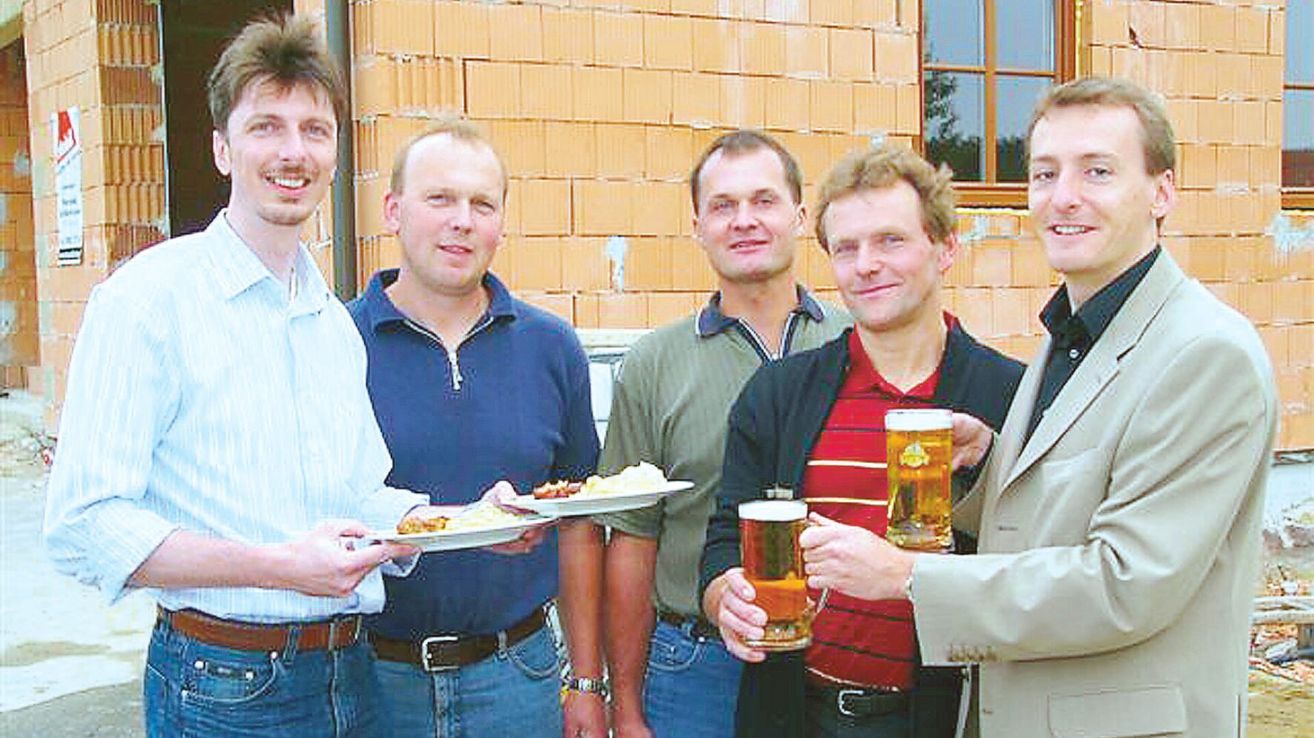 Fünf Männer stehen lächelnd vor einem Ziegelgebäude, halten Teller mit Essen und Bierkrügen in den Händen.