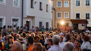 Eine Menschenmenge versammelt sich in einer städtischen Umgebung, klatscht und lächelt vor Gebäuden mit Bannern und Blumen.