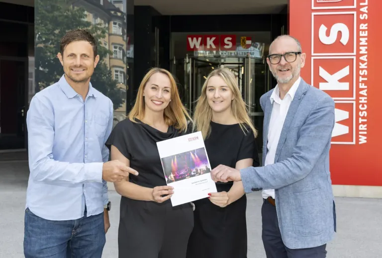 Zwei Frauen und zwei Männer stehen lächelnd vor einem Gebäude und halten ein Zertifikat in der Hand. Die Frauen tragen schwarze Kleider und die Männer hellblaue und dunkelblaue Anzüge. Hinter ihnen hat das Gebäude eine rote Wand mit den Buchstaben 'WKS' und einem Logo.