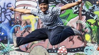 Ein Mann in einem karierten Hemd und Jeans springt vor einer farbenfrohen Wandmalerei an einer Wand in die Luft.
