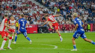 Ein Fußballspieler in einem roten Trikot mit der Nummer 27 springt in die Luft, während ein anderer Spieler in einem blauen Trikot zusieht.
