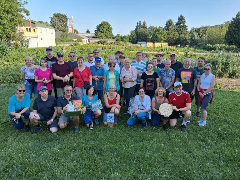 Eine Gruppe von Menschen posiert für ein Foto im Garten, einige halten Geschenke und ein Mann hält eine runde Plakette. Hinter ihnen ist ein Feld mit Bäumen und Gebäuden.