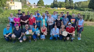 Eine Gruppe von Menschen posiert für ein Foto im Garten, einige halten Geschenke und ein Mann hält eine runde Plakette. Hinter ihnen ist ein Feld mit Bäumen und Gebäuden.