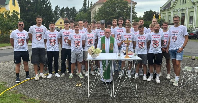 Eine Gruppe von Fußballspielern steht mit einem Priester vor einem Tisch mit einem Kreuz und Kerzen. Bäume und Gebäude sind im Hintergrund.
