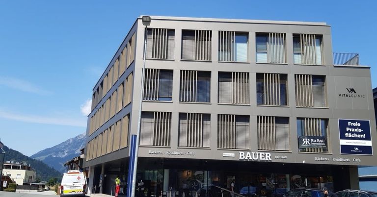 Ein modernes Gebäude mit Glasfenstern und einem blauen Vordach, auf dem in großen weißen Buchstaben der Schriftzug 'Bauer' steht. Davor befindet sich eine Straßenlaterne und ein geparktes Fahrzeug.