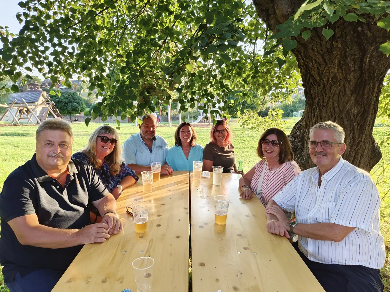 Eine Gruppe von Menschen sitzt um einen Tisch mit Biergläsern herum, einige mit Sonnenbrille, unter einem Baum in einem Park.