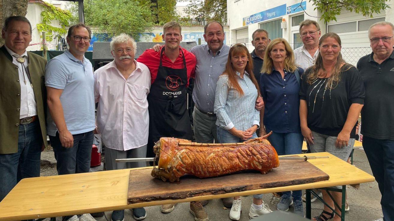 Eine Gruppe von Menschen, darunter ein Mann in einer Schürze, steht um ein großes gebratenes Schwein auf einem Holztisch. Sie lächeln und scheinen für ein Foto zu posieren.