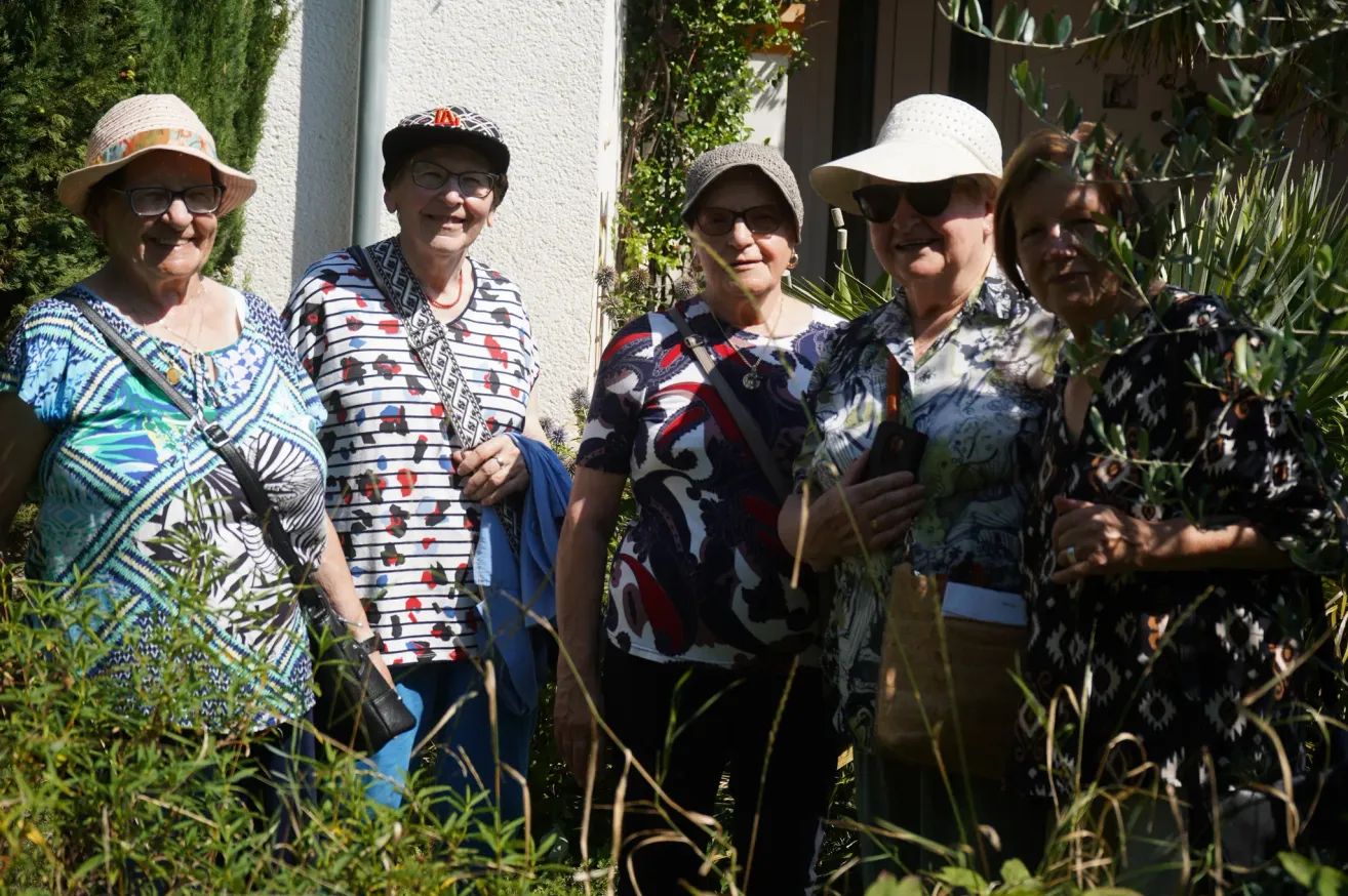 Fünf Frauen, jede mit Hut und Sonnenbrille, stehen dicht beieinander in der Nähe eines Gebäudes. Sie lächeln und scheinen für ein Foto zu posieren.