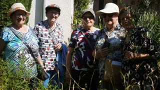 Fünf Frauen, jede mit Hut und Sonnenbrille, stehen dicht beieinander in der Nähe eines Gebäudes. Sie lächeln und scheinen für ein Foto zu posieren.