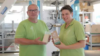 Zwei Personen in grünen Hemden stehen in einer Fabrik. Der Mann hält eine Tüte mit Körnern und die Frau hält eine Schachtel Reis.