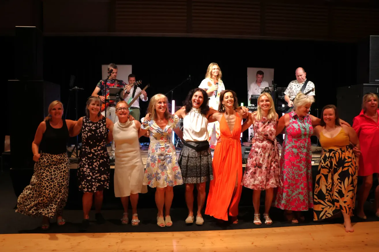 Eine Gruppe von Frauen steht auf der Bühne mit einer Band hinter ihnen, lächelt und posiert für ein Foto. Die Frauen sind in bunten Kleidern gekleidet. Die Bandmitglieder spielen Gitarren und andere Instrumente.