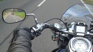 Ein Motorradfahrer fährt auf einer Straße, mit klarem Blick auf den Tachometer und die Lenker. Der Fahrer trägt Handschuhe und das Motorrad hat eine Windschutzscheibe.