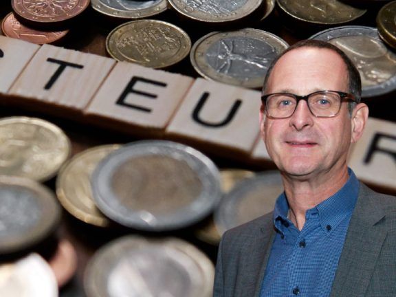Ein Mann in Anzug und Brille steht vor einem Haufen Münzen, auf denen das Wort 'TEU' in Scrabble-Steinen geschrieben ist.