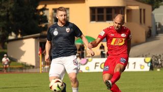 Zwei Fußballspieler in Trikots spielen auf dem Feld. Der Spieler in Schwarz versucht, dem Spieler in Rot den Ball abzunehmen.