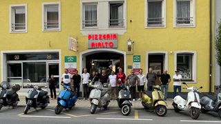 Eine Gruppe von Menschen steht vor einem gelben Gebäude mit einem Schild, das 'Pizzeria Rialto' liest. Sie posieren mit ihren Rollern vor dem Restaurant.
