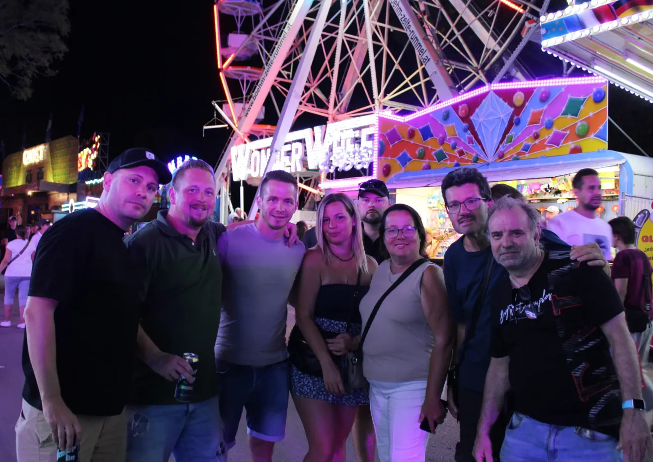 Eine Gruppe von Menschen steht bei Nacht vor einem leuchtenden Karussell. Sie lächeln für die Kamera.