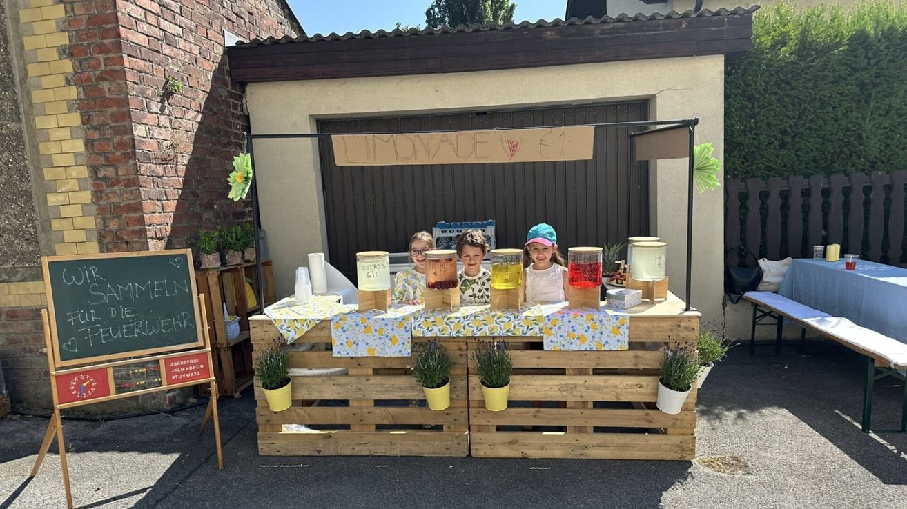 Drei Kinder stehen hinter einem Limonadenstand, lächeln und posieren für ein Foto. Eine Holzstruktur hält Gläser mit Limonade und Zitronen. Der Stand ist mit gelben Tischdecken und Topfpflanzen dekoriert.