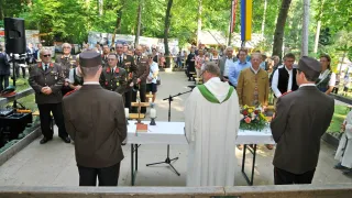 Eine feierliche Veranstaltung findet im Freien statt, angeführt von einem Priester, umgeben von einer Menschenmenge. Der Priester trägt eine weiße Robe und einen grünen Umhang. Vor ihm steht ein Tisch mit einem Buch, Kerzen und Blumen.