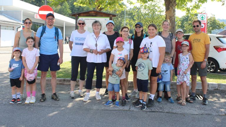 Eine Gruppe von Menschen, einschließlich Erwachsener und Kindern, posiert für ein Foto in einem Park. Sie stehen vor einem Holzschild und Bäumen.