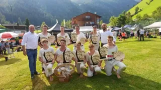 Eine Gruppe junger Männer in weißen Uniformen posiert vor einem Berghintergrund für ein Foto und hält Holzplaketten.