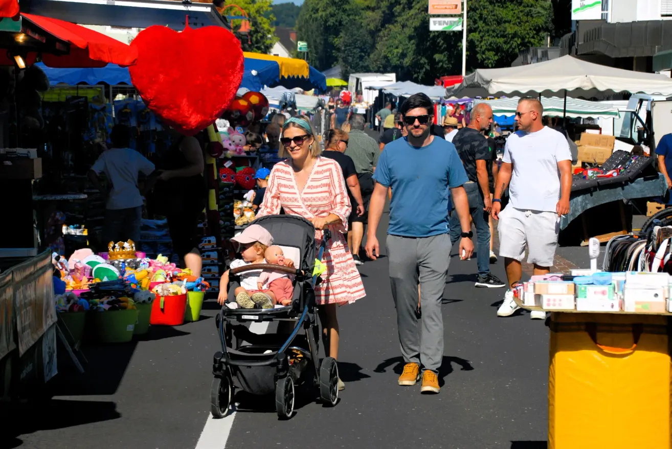 Ein Paar mit einem Baby im Kinderwagen geht eine belebte Straße auf einem Markt entlang, mit einem riesigen roten Herzdekoration über ihnen.